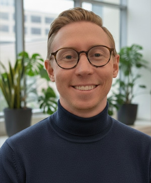 Staff portrait of Benjamin Morse. Wearing a navy turtleneck sweater poses indoors in front of large windows, with several green potted plants visible in the background.