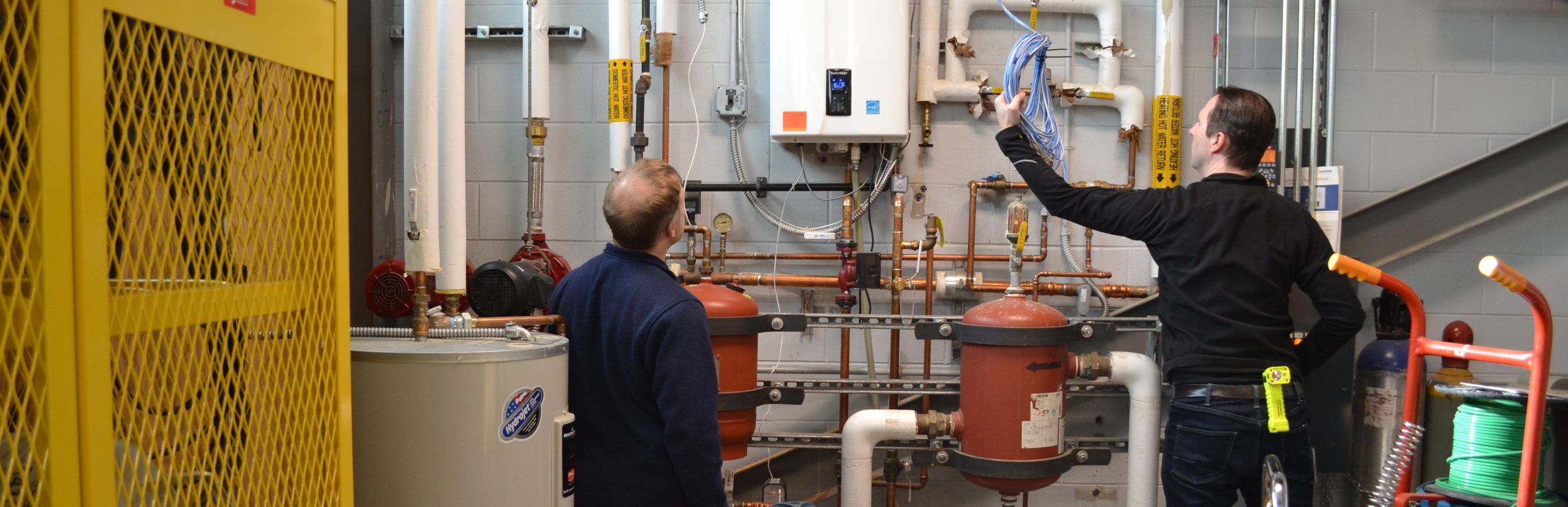 Two individuals are standing in a mechanical room filled with pipes, tanks, and industrial equipment. One person is holding a bundle of blue network cables near an electrical panel, while the other observes the setup. Various insulated pipes, copper tubing, and red cylindrical tanks are visible along the wall, along with a yellow metal safety cabinet on the left and a spool of green wire on a cart to the right. The environment suggests facility maintenance or system inspection work.
