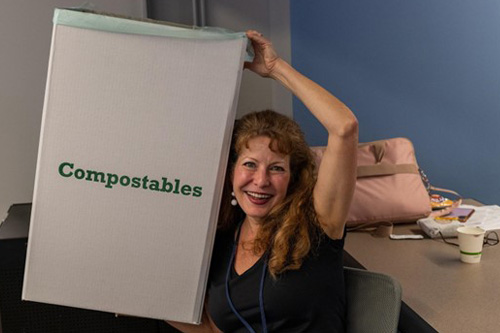 A person holding up a large white bin labeled "Compostables" in green text, sitting at a desk with a pink bag, paper cup, and various items in the background.