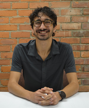 Staff portrait Sam Kocurek - gentleman wearing a dark grey shirt sitting at a white table.